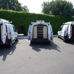 Rear view of Fleet of three white Ford vans with roof-mounted tube carriers and interior shelving.