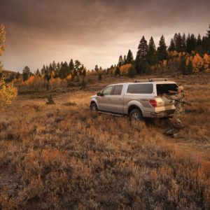 Ford F-150 recreational truck with Leer 100XQ camper shell, hunter in camouflage opening the rear hatch, parked off-road in fall-colored backcountry.