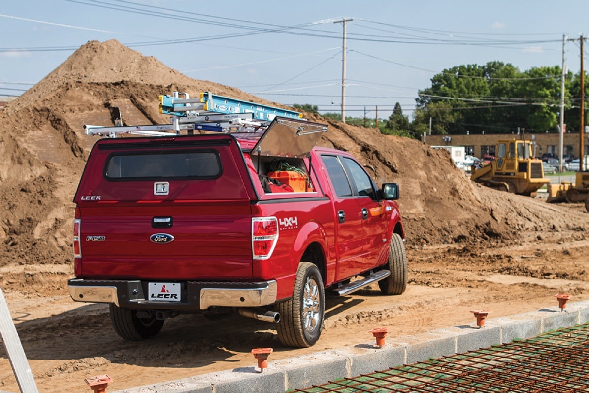 Work truck from our work trucks and vans lineup with red SnugTop camper shell, side panel open, and overhead ladder rack at a construction site.