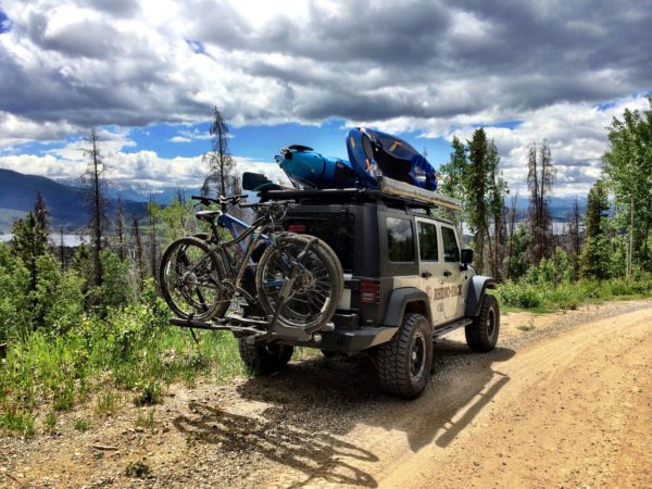 Adventure-ready Jeep with Rhino Rack carrying kayaks on the roof and mountain bikes on the back, driving a scenic mountain dirt road near trees, lake, and clouds.