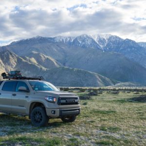 Toyota Tacoma off-road recreational truck with soft camper shell and Rhino Rack gear system, parked in grassy valley with mountain backdrop.