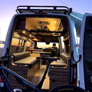 Lifestyle adventure van with bike carrier, window shades, surfboard mount, sink, swivel table, and seating.