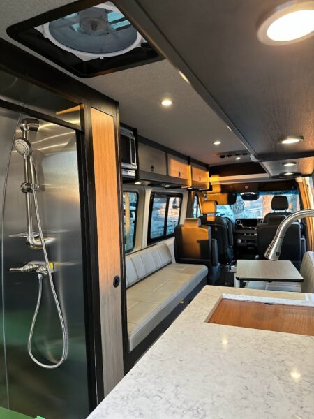 Custom travel van interior showing shower, roof vent, seating and counter space with sink.