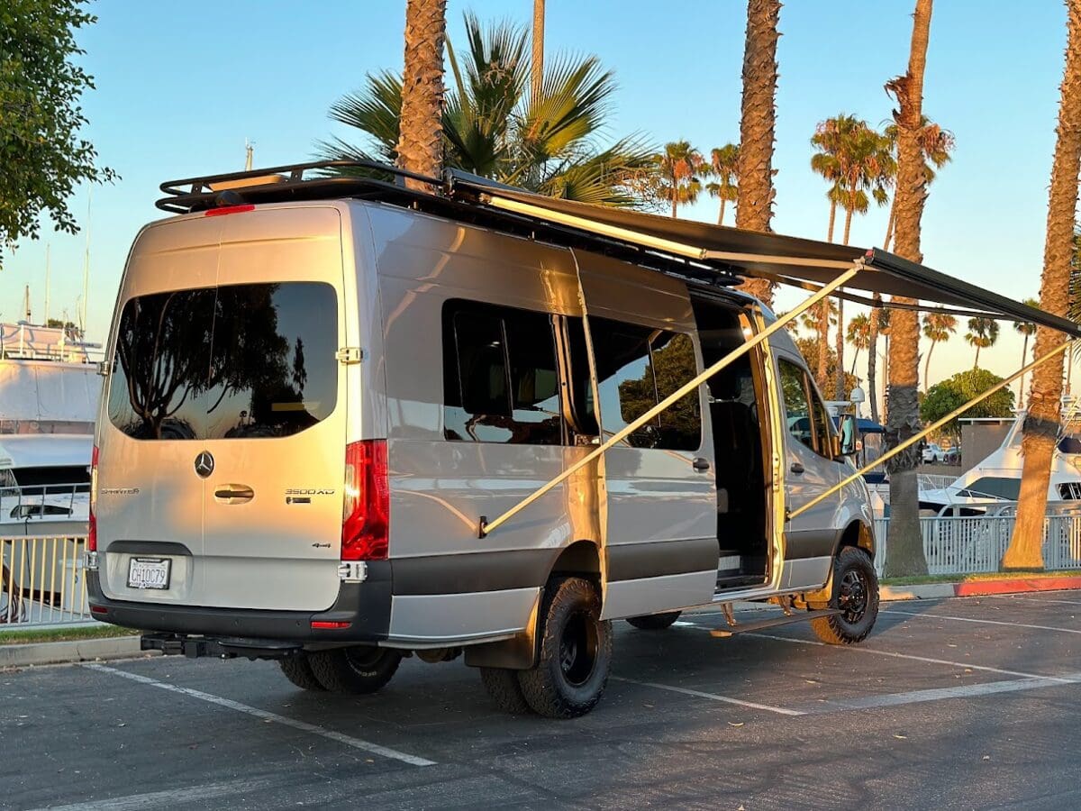 Upfitted Mercedes 3500 AD 4x4 van with dually tires, roof rack, and side awning by the marina.